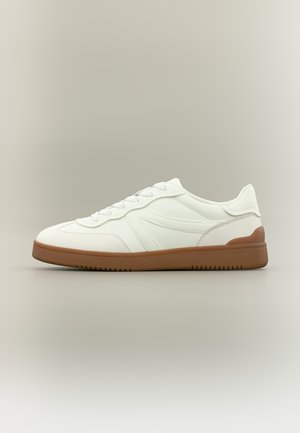 Witte lage sneaker met witte veters en bruine rubberen zool, zijaanzicht op een neutrale grijze achtergrond.