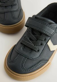 Baskets noires avec une semelle gomme texturée, dotées de lacets et d'une sangle en Velcro. Deux chaussures sont présentées sur un fond clair.