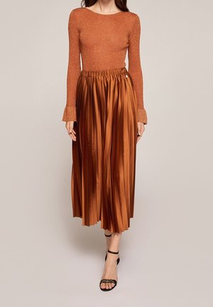 Vrouw draagt een nauwsluitende, roestkleurige top met lange mouwen en een high-waisted, geplooide metallic koperen midi-rok met zwarte enkellaarzen met riempjes.