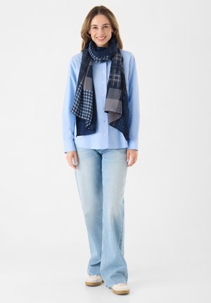 Chemise bleu à boutons et manches longues, associée à un jean évasé bleu clair et à une écharpe en navy à motifs multiples. Des chaussures décontractées complètent le look.