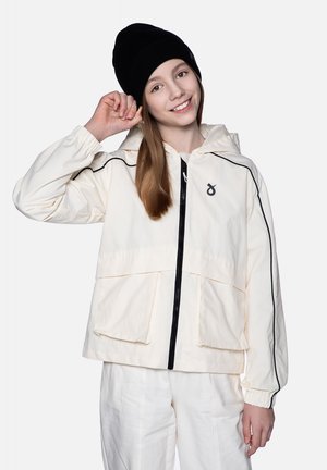 Kremowa kurtka z kapturem z czarnymi akcentami, z przodu na zamek, z dużymi kieszeniami. Model nosi czapkę beanie w kolorze czarnym i białe spodnie.
