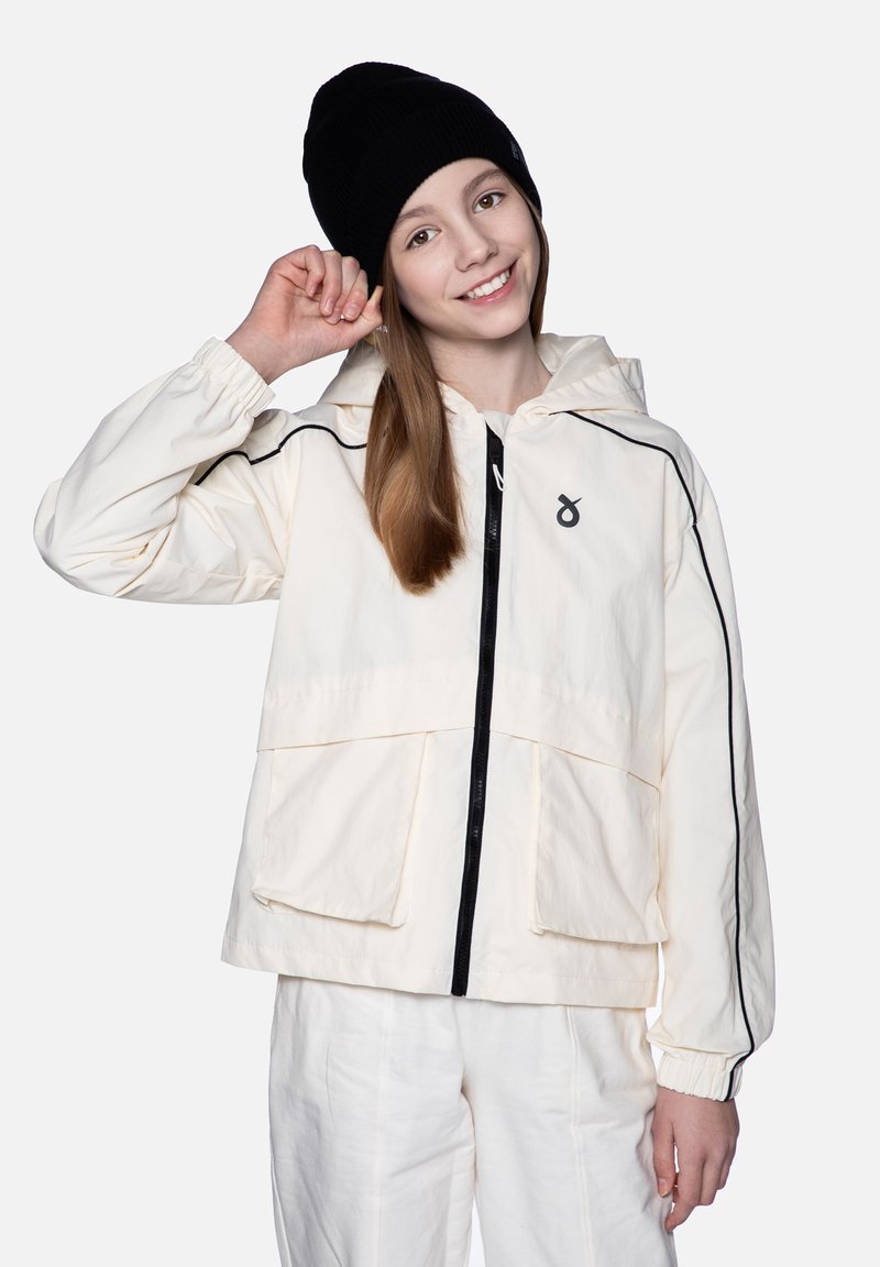 Kremowa kurtka z kapturem z czarnymi akcentami, z przodu na zamek, z dużymi kieszeniami. Model nosi czapkę beanie w kolorze czarnym i białe spodnie.