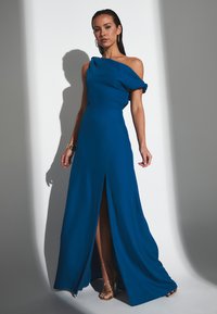 Blaue Abendkleid mit einem einzelnen schulterfreien Träger, seitlichem Schlitz, fließendem Stoff und subtiler Glanz. Kombiniert mit metallischen Riemchen-Heels.