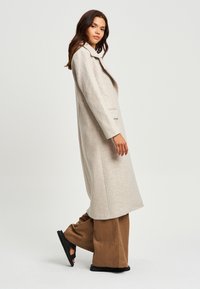 Cappotto lungo beige in lana con colletto a punte, tasche laterali e taglio dritto, abbinato a pantaloni a gamba larga marroni e sandali neri.