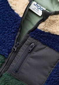 Fleece bunda s modrými, zelenými a béžovými panelmi, s prednou zipsovou kapsou, textúrovanými fleece sekciami a štítkom označujúcim veľkosť 12-18 mesiacov.