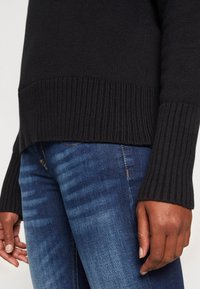 Personne portant un pull côtelé sombre et un jean bleu délavé, debout avec les mains détendues le long du corps.