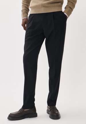 Pantalones negros de corte recto, con pliegues en la parte delantera, confeccionados en una tela suave. Llevados con mocasines marrones.