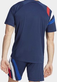 Man som bär en marinblå sporttröja och shorts med röd, vit och blå rand, sedd bakifrån mot en enkel bakgrund.