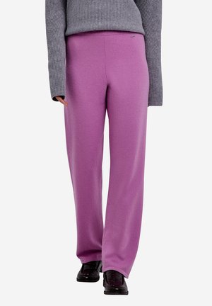 Person trägt lockere lila Hose, grauen langärmeligen Pullover und schwarze Schuhe, steht vor einfachem Hintergrund.