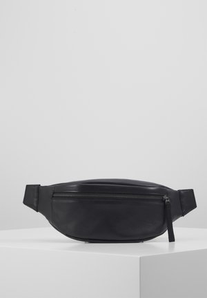 Sac banane en cuir noir avec poche zippée à l'avant, présenté sur une surface blanche sur fond gris uni.