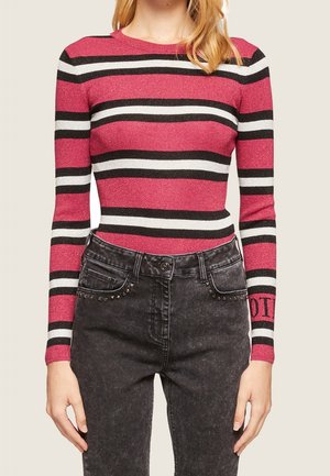 Kvinde iført en tætsiddende langærmet skjorte med lyserøde, sorte og hvide horisontale striber kombineret med sort high-waist jeans med nitte detaljer.