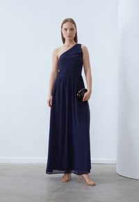 Jūras spēku viens plecu maxi kleita ar pielāgotu augšdaļu un plūstošu svārku, kurai ir savilkts jostasvieta un caurspīdīga tekstūra. Modelis tur melnu clutch.