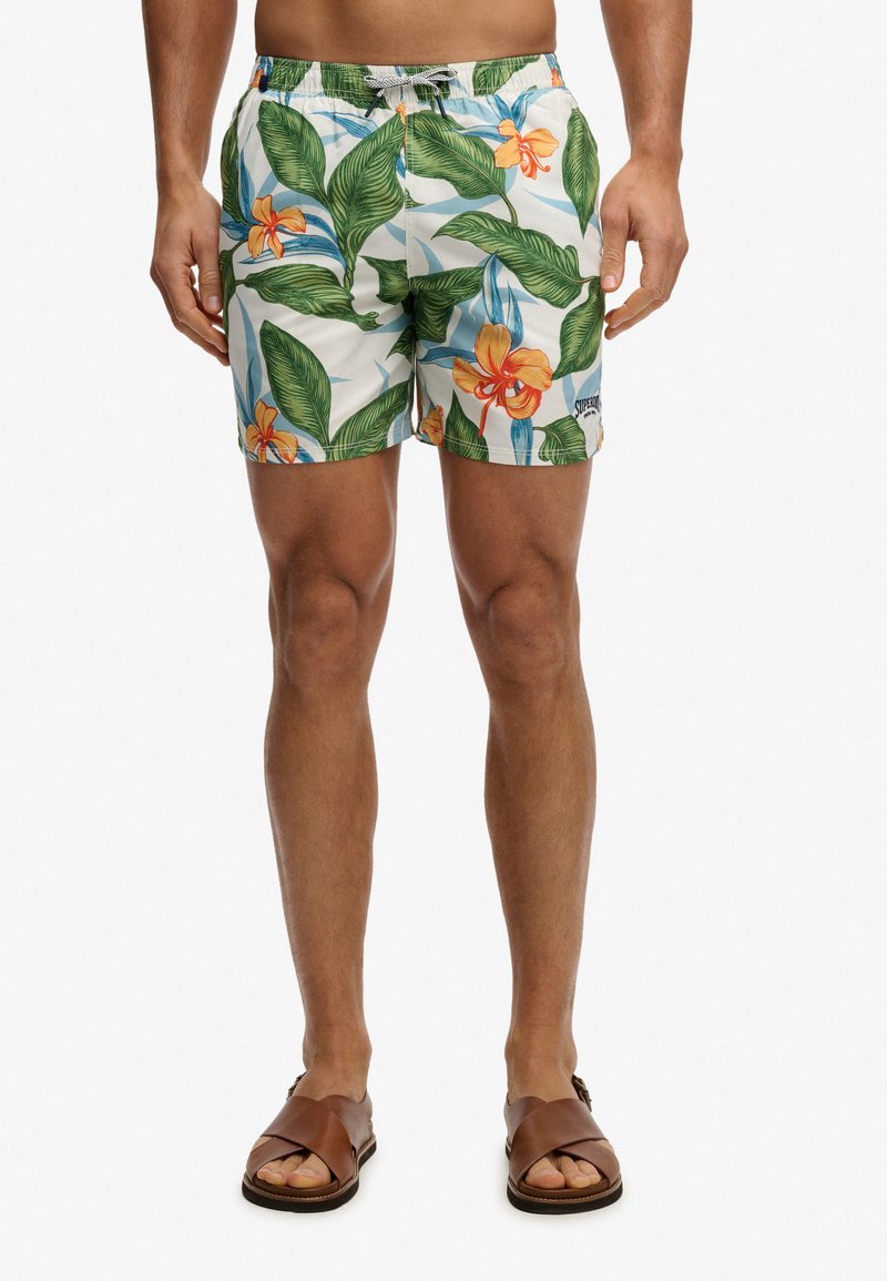 Homme portant un short de bain à imprimé floral avec des feuilles vertes et des fleurs orange, ainsi que des sandales en cuir marron, debout devant un fond blanc.