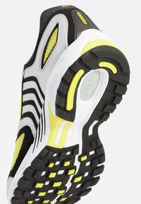 Sapatilha com parte superior em preto, cinza e branco, apresentando detalhes em amarelo. Sola texturizada com um padrão de pega proeminente e detalhes de marca.