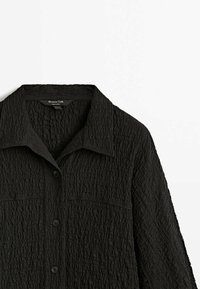 Chemise noire texturée à boutons, avec col, manches longues et deux poches poitrine.