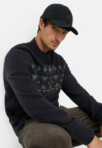 Marineblauer Strickpullover mit einem geometrischen Muster in Blau, Braun und Creme. Das Modell trägt zudem eine schwarze Mütze. Cordhosen in einem gedämpften Farbton.