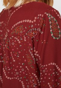 Blouse bordeaux avec un motif floral, présentant de petites fleurs roses, blanches et vertes, et une coupe ample avec un dos froncé.