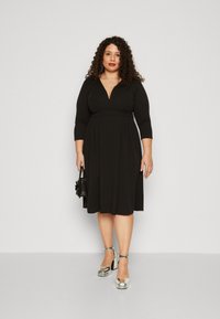 Vestido negro de longitud a la rodilla con escote en V, mangas tres cuartos y pliegues en la cintura. La modelo lleva tacones plateados gruesos y sostiene un bolso negro.