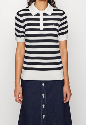 Vrouw die een zwart-wit horizontaal gestreept poloshirt met korte mouwen draagt en een donkerdenim rok met knopen aan de voorkant.