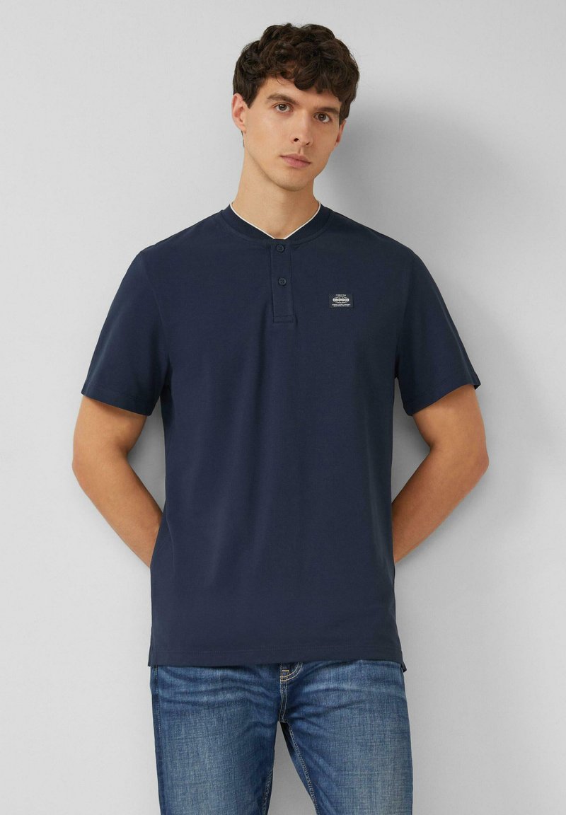 Jeune homme aux cheveux bouclés portant un t-shirt henley à manches courtes bleu marine et un jean bleu, debout les mains derrière le dos.
