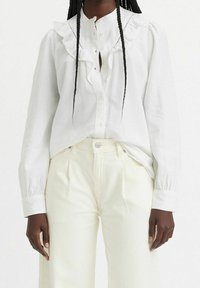 Vit skjorta med volangkrage och manschetter, i kombination med krämfärgade högmidjade byxor. Slät tyg med subtil lyster.