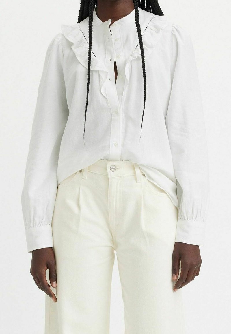 Vit skjorta med volangkrage och manschetter, i kombination med krämfärgade högmidjade byxor. Slät tyg med subtil lyster.