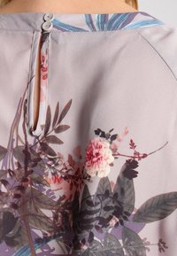 Blomsterprintstof med en lys lilla baggrund. Har en nøglehulåbning bagpå, sikret med to hvide knapper.