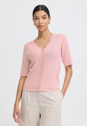 Femme portant un haut en maille rose à manches courtes boutonné avec des bords festonnés et un pantalon de couleur crème, debout avec une main dans la poche.