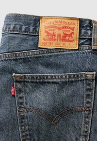 Jeans i denim med en ljusblå tvätt, med orange sömmar, en läderetikett som visar märke och modell, samt en röd logotabby på bakfickan.