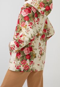Persona vistiendo una sudadera crema con patrones florales rojos, rosas y amarillos, combinada con pantalones marrón claro, con las manos en los bolsillos, vista de perfil.