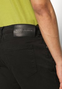 Černé džíny s koženým štítkem na pase s nápisem "BALDESSARINI JEANS." Vyznačují se výrazným prošíváním a detaily na zadních kapsách.