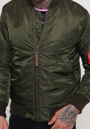 Mann trägt eine olivgrüne Bomberjacke mit gerippten Bündchen und Bund, mit Reißverschluss vorne und einem kleinen roten Etikett an der linken Ärmeltasche.