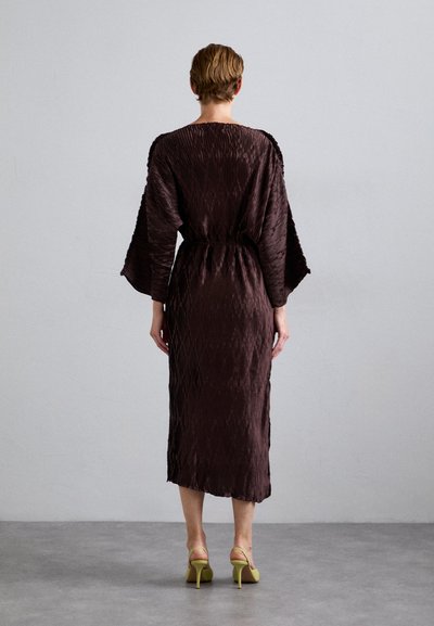 Person standing facing away wearing a dark brown textured midi dress with wide sleeves and yellow high-heeled shoes on a gray floor.
