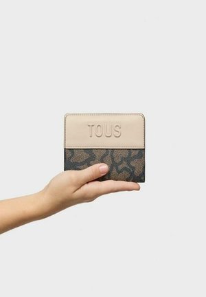 Cartera en crema y marrón oscuro de Tous, con un panel superior liso y una sección inferior texturizada con un patrón sutil, sostenida en una mano.