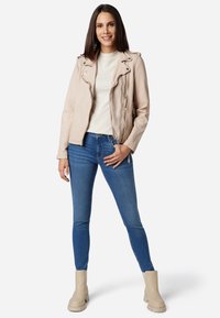 Ljus beige imitation läder motorcykeljacka med dragkedjefickor, parat med blå skinny jeans och beige ankelstövlar, mot en vit bakgrund.