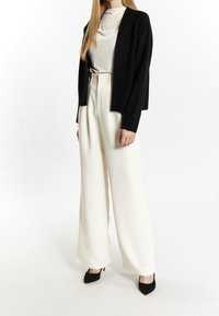 Schwarzer Cardigan über einem hellbeigen Top, kombiniert mit hochgeschnittenen, weit geschnittenen cremefarbenen Hosen. Schwarze spitz zulaufende High Heels vervollständigen das Outfit.