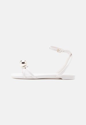Sandale plate blanche avec fines lanières, boucle à la cheville et un grand détail boule argentée brillante à l'avant, au niveau des orteils.