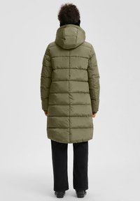 Osoba u dugom maslinasto zelenom puffer kaputu s kapuljačom, crnim hlačama i crnim cipelama, stoji okrenuta leđima prema jednostavnom pozadinskom zidu.