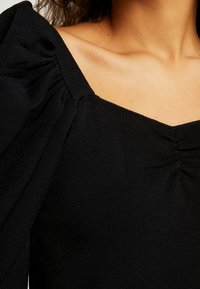 Zwarte top met een vierkante halslijn en geplooide pofmouwen. De textuurstof is geribbeld, met subtiele stiksels en een getailleerde silhoette.