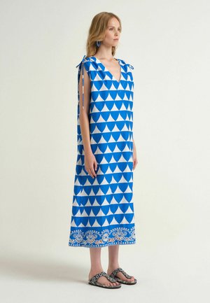 Donna che indossa un vestito smanicato con motivo geometrico blu e bianco, con spalline annodate e sandali neri con perline decorative.