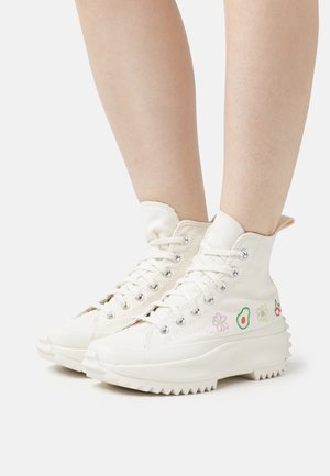 Witte hoge sneakers met dikke zolen, geborduurd met kleurrijke bloemen, een avocado en kersen, gedragen op blote benen tegen een effen achtergrond.