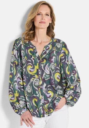 Vrouw met blond haar, gekleed in een knoopblouse met lange mouwen in een abstracte paisleypatroon in groen, geel, paars en wit, en witte broek.