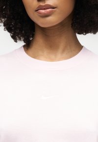 Κοντινό πλάνο στο κάτω μέρος του προσώπου και στον λαιμό γυναίκας με σγουρά μαλλιά, που φοράει ένα ανοιχτό ροζ μπλουζάκι Nike με λευκό λογότυπο.