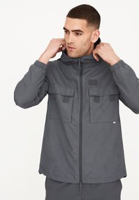 Graue leichte Jacke mit Reißverschluss vorne, zwei Brusttaschen, elastischen Bündchen und einer Kapuze. Glatte Textur mit minimalen Brandingdetails.