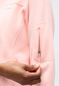 Χέρι που τραβά το φερμουάρ στην τσέπη του πάνω βραχίονα ενός ροζ φλις μπουφάν, δείχνοντας την υφή του υφάσματος και τις λεπτομέρειες του φερμουάρ.