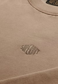 Sweatshirt marron clair en tissu texturé avec un logo "Superdry" brodé discret sur la poitrine. Col rond.