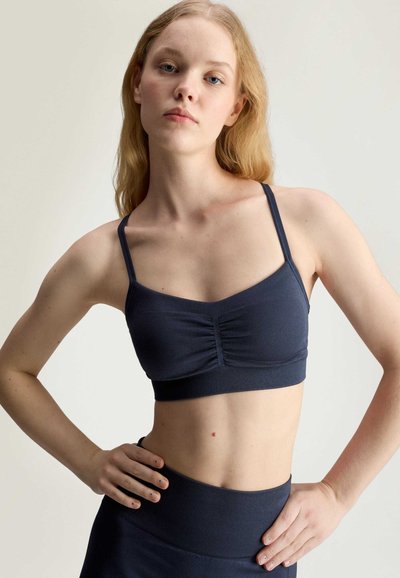Young woman with long blonde hair wearing a navy blue sports bra and high-waisted leggings, standing with hands on hips against neutral background.