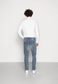 Camisa blanca de manga larga combinada con vaqueros de mezclilla azul claro. Los vaqueros tienen un lavado desgastado y detalles de costura sutiles. Zapatillas blancas completan el conjunto.
