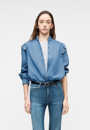 Donna che indossa una camicia di jeans con spalle arricciate blu sopra una maglietta bianca, infilata in jeans skinny blu a vita alta con una cintura nera.