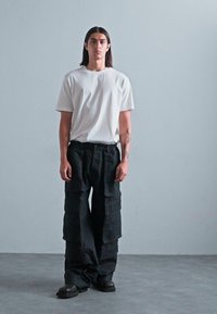 Young person standing, wearing a white t-shirt, loose black cargo pants, and black boots against a plain gray background.